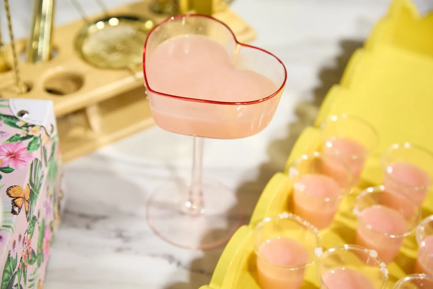Pink cocktail in a heart-shaped coupe glass beside a tray of pink shot samples
