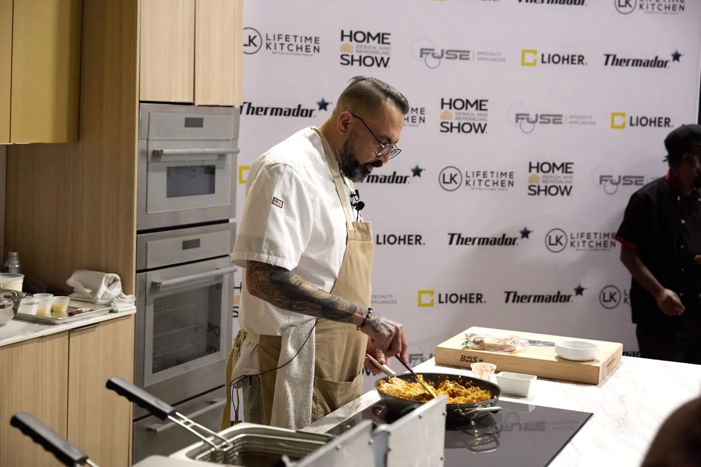 Chef cooks in a skillet at a demo kitchen island in front of a sponsor step-and-repeat backdrop