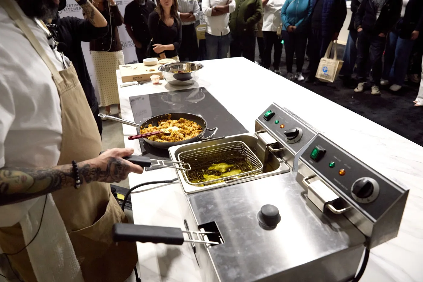 Chef fries samosas in a tabletop deep fryer while a crowd watches at the cooking demo