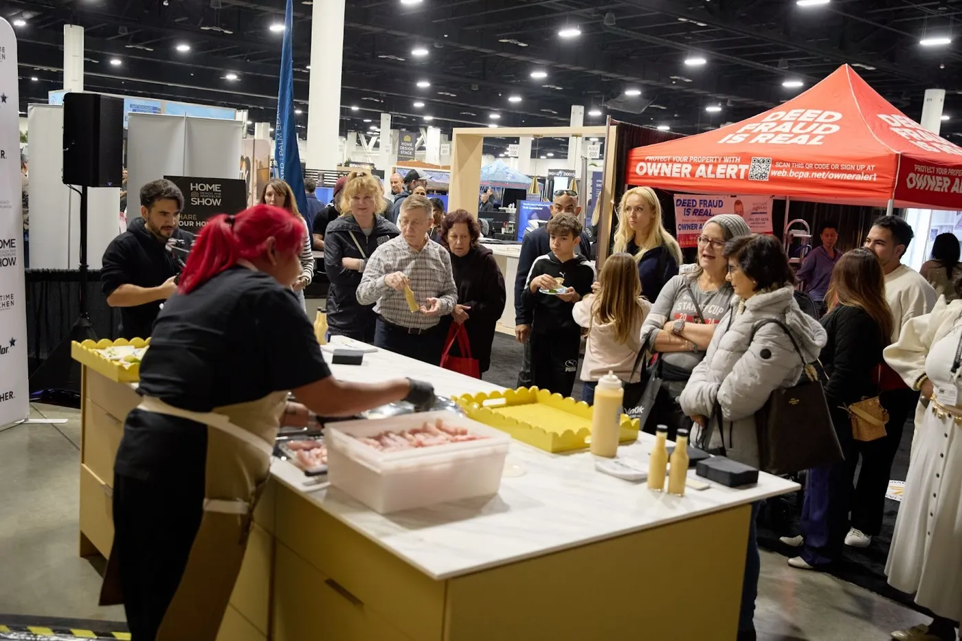 Crowd gathered around a chef's cooking demo booth at the Home Show, near a Deed Fraud Owner Alert tent