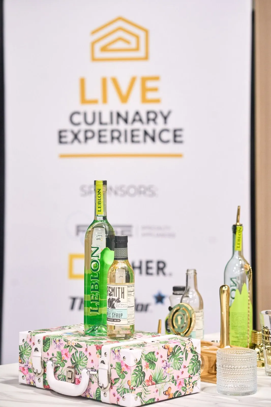 Leblon cachaca and maple syrup bottles on a floral suitcase bar cart at a Live Culinary Experience booth