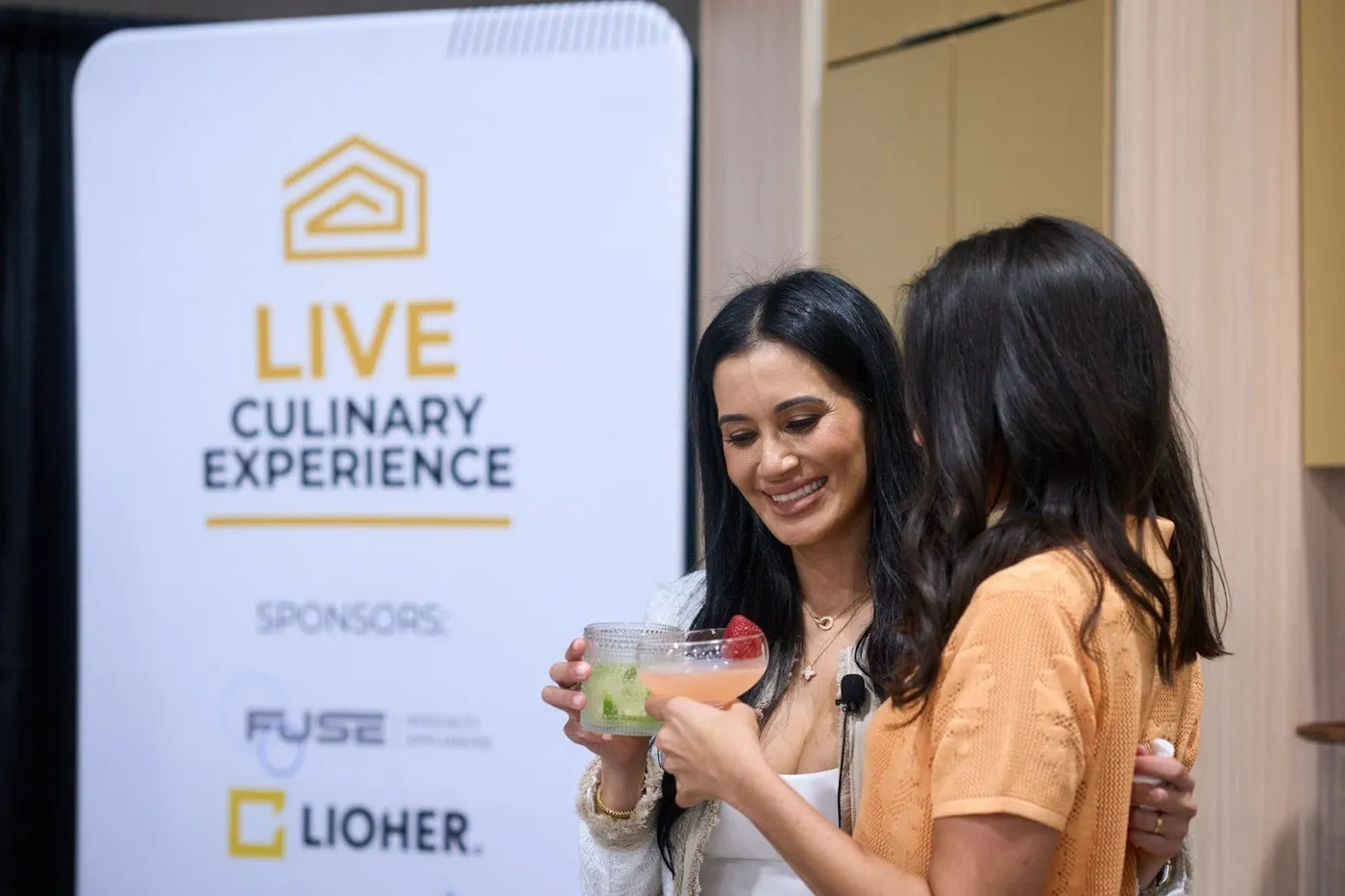 Two women toasting with cocktails beside a Live Culinary Experience sponsor sign