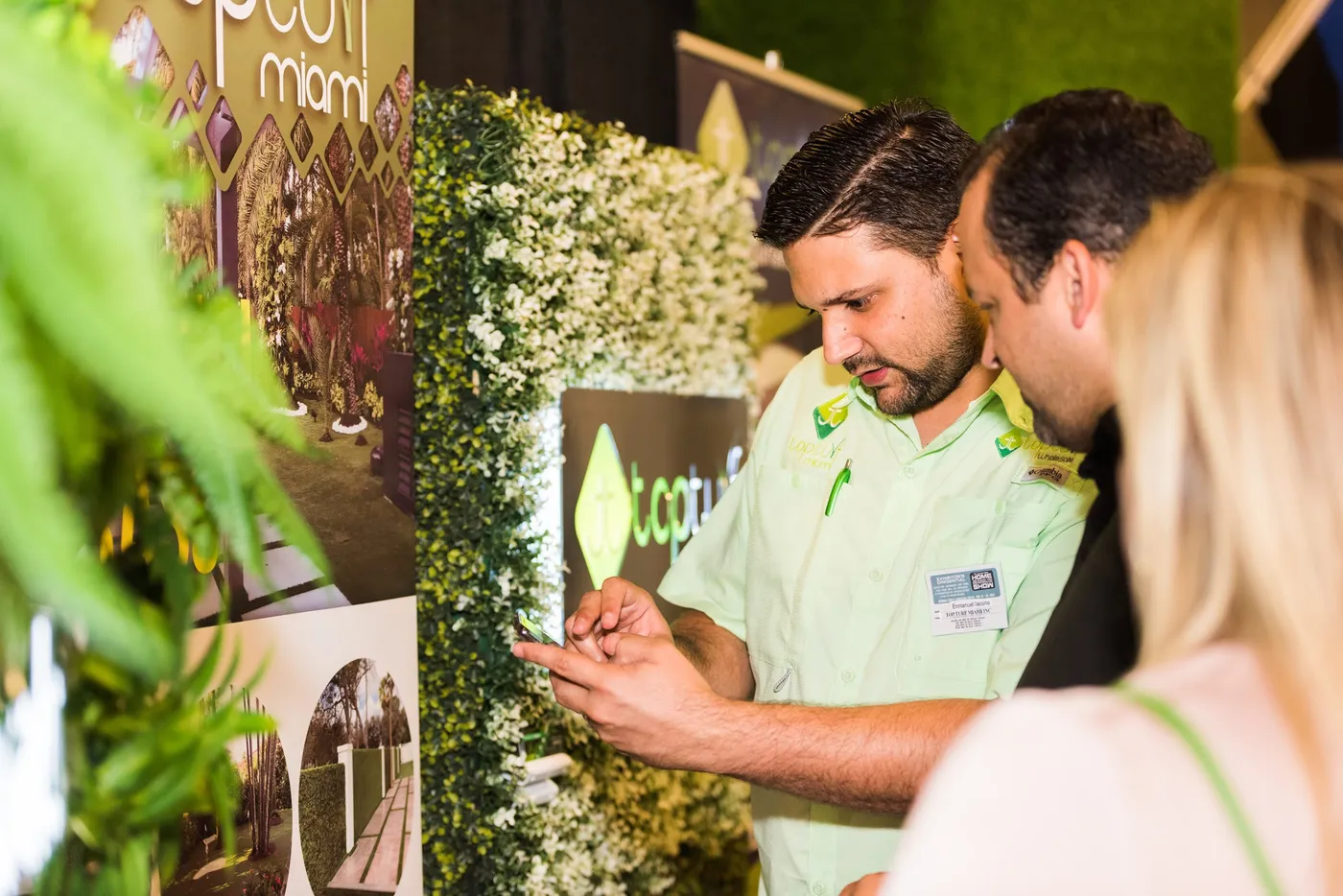 Attendees viewing a phone at the Topturf Miami artificial turf booth with a green foliage display