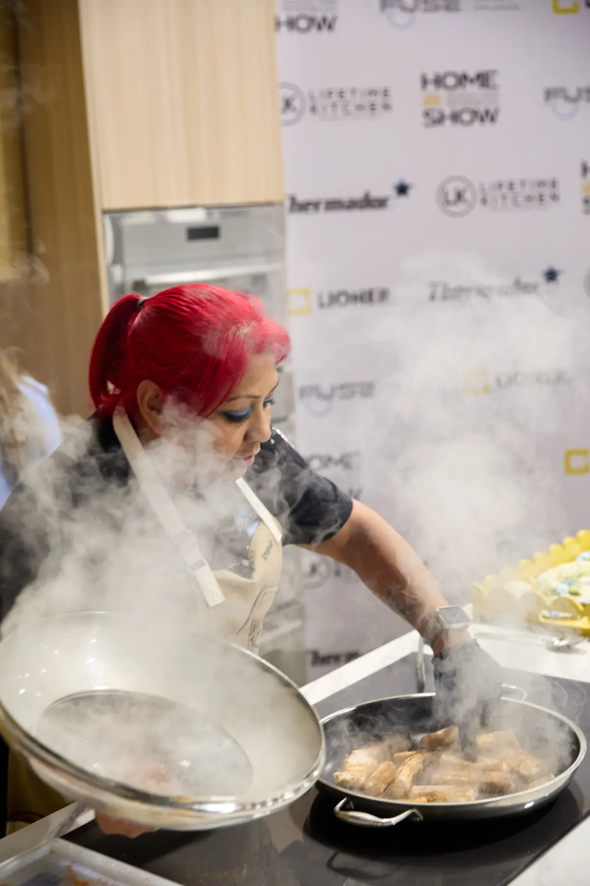 Chef in Home Show apron cooks over billowing steam on the culinary stage, Home Show branded backdrop behind her
