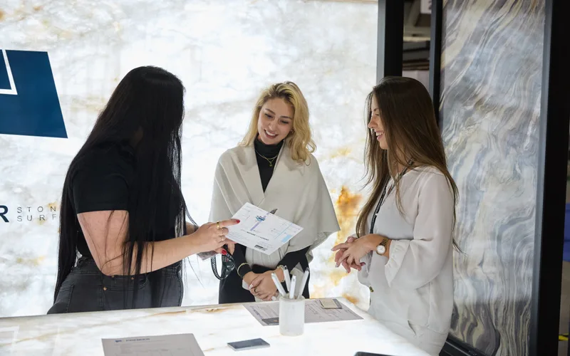 Three women laugh and review a brochure at a stone surfaces exhibitor booth